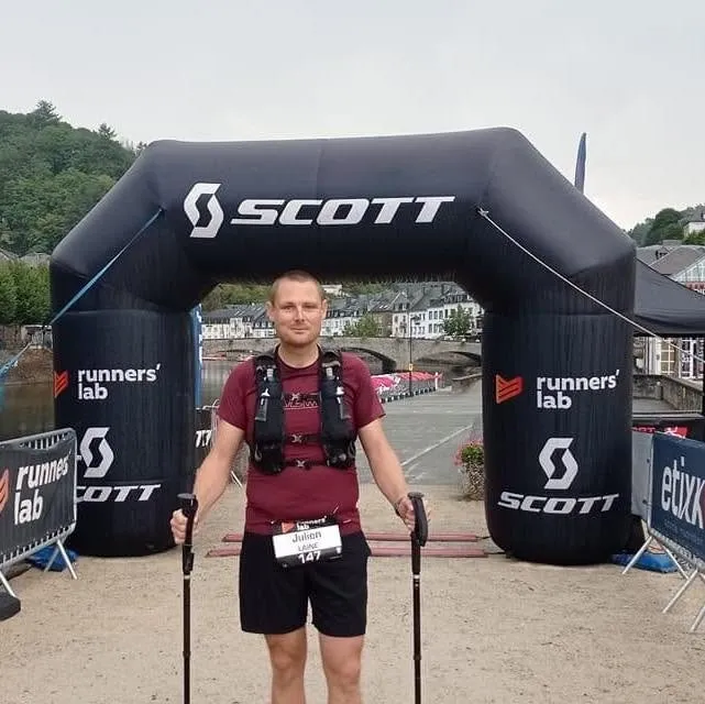 Julien Lainé sous l’arche d’arrivée d’un trail, dossard au nom de Julien Lainé, bâtons en main