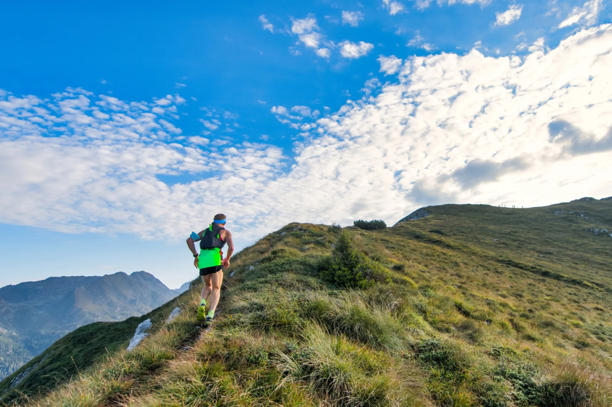 Traileur en montée sur un sentier de crête herbeux, sous un ciel bleu chargé de nuages